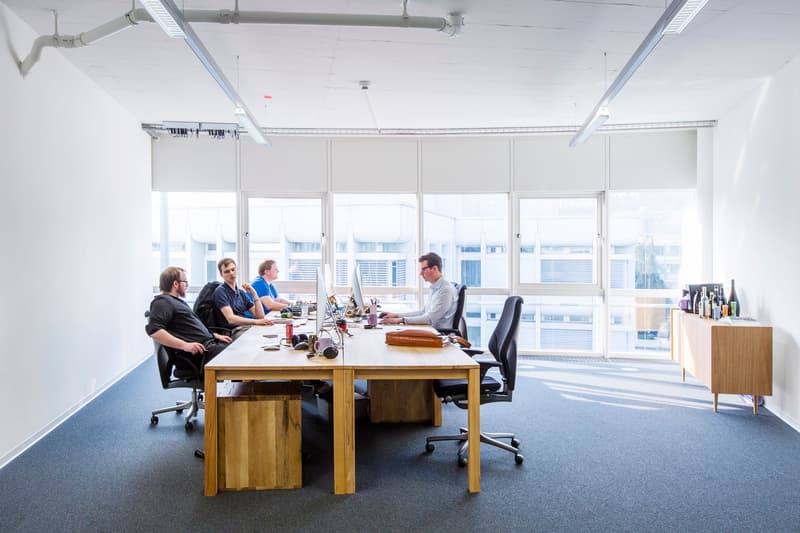 Open office space with large windows, wooden desks, office chairs, and various office equipment and supplies