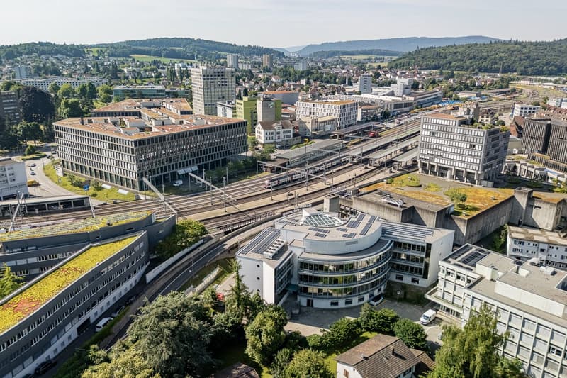 Aerial view of a city skyline with a mix of modern high-rise buildings and older structures. The city appears to be situated in a hilly, forested area with a train station or transportation hub visible in the center of the image. There are several parking