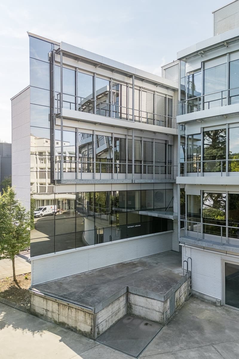 Multi-story commercial or office building with a modern, glass-and-steel facade. The building has multiple levels, with balconies or terraces visible on some floors. The ground floor appears to have a raised entrance platform or steps leading up to the ma