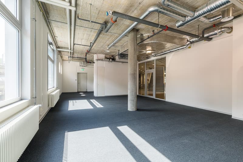 Large open office space with concrete ceiling, exposed pipes and ductwork, gray carpeted floor, white walls, and a door leading to another area.