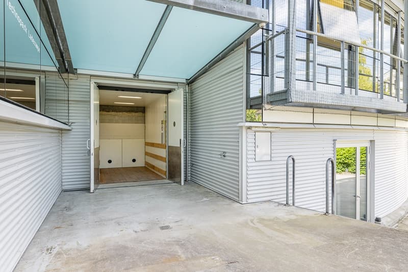 The image shows a storage unit or garage space with a large open doorway. The space has a concrete floor and corrugated metal walls and ceiling. There are some boxes or storage containers visible inside the unit.