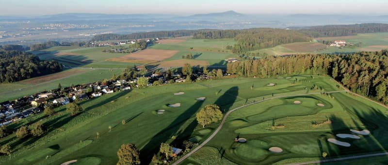 Aerial view of a golf course, wide grass fairways, green bushes, trees, a residential area in the distance, clear sky
