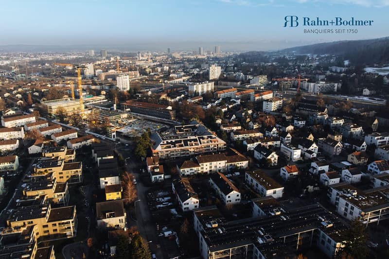 An aerial view of a city with many buildings, including some with solar panels on the roofs, in the foreground. The city has an extensive layout with buildings of different sizes and designs. Some of the buildings have cranes on top, suggesting ongoing co