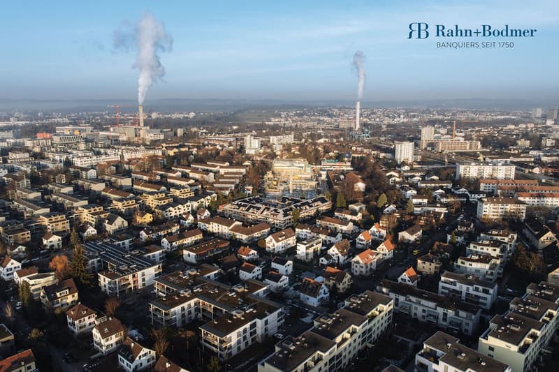 aerial view of a city with various buildings and structures, some emitting smoke, surrounded by trees and roads, with industrial areas in the distance
