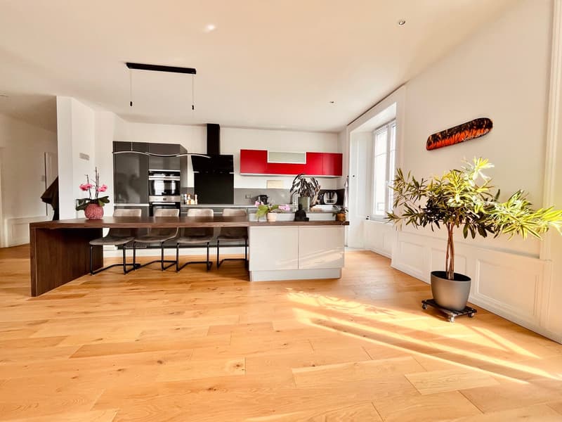 Modern kitchen with wooden dining table, white cabinets, red appliances, central island with plants, large windows, hardwood floors.