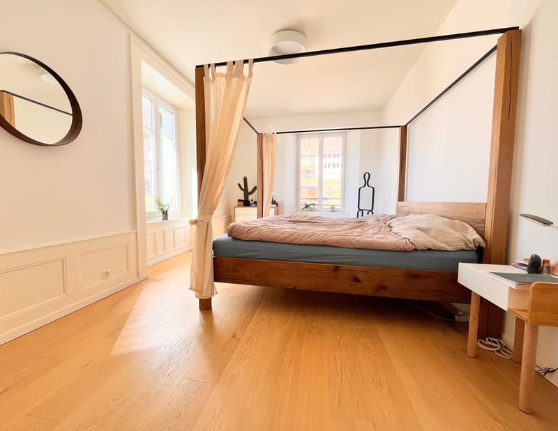 wooden canopy bed, blue mattress, white ceiling lamp, large round mirror, wooden desk, plants on the window sill
