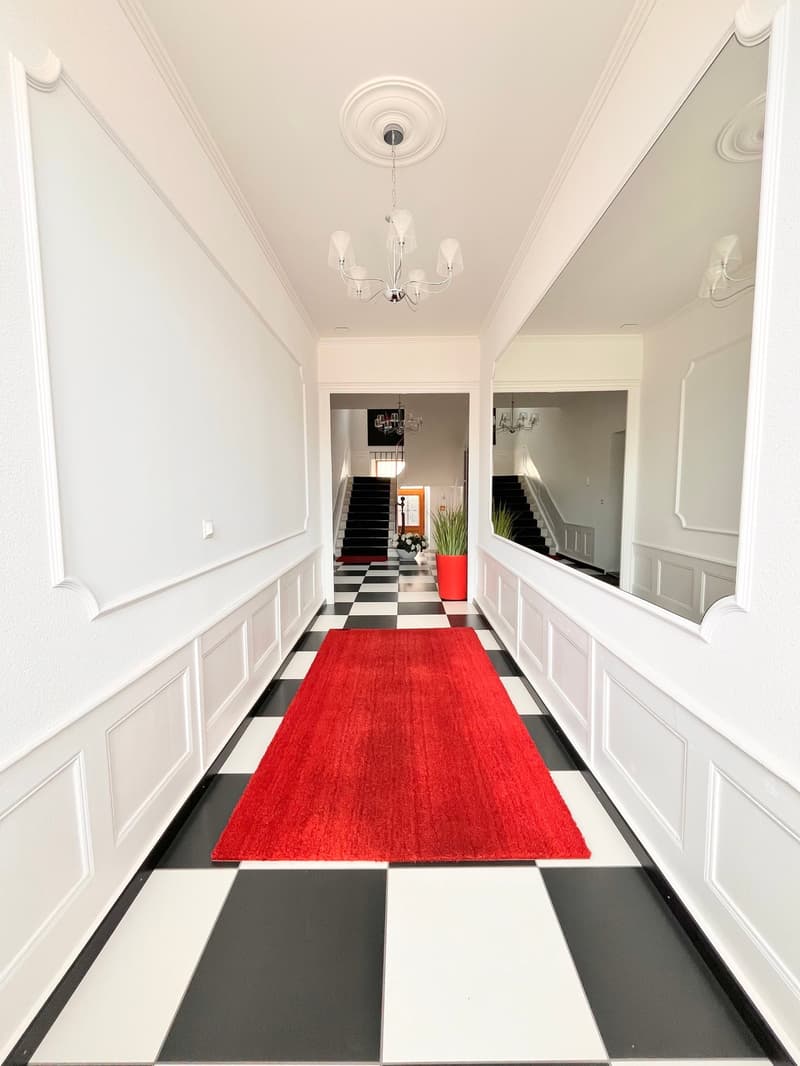 long hallway, white walls, black and white checkered floor, red carpet, chandelier, mirror, staircase