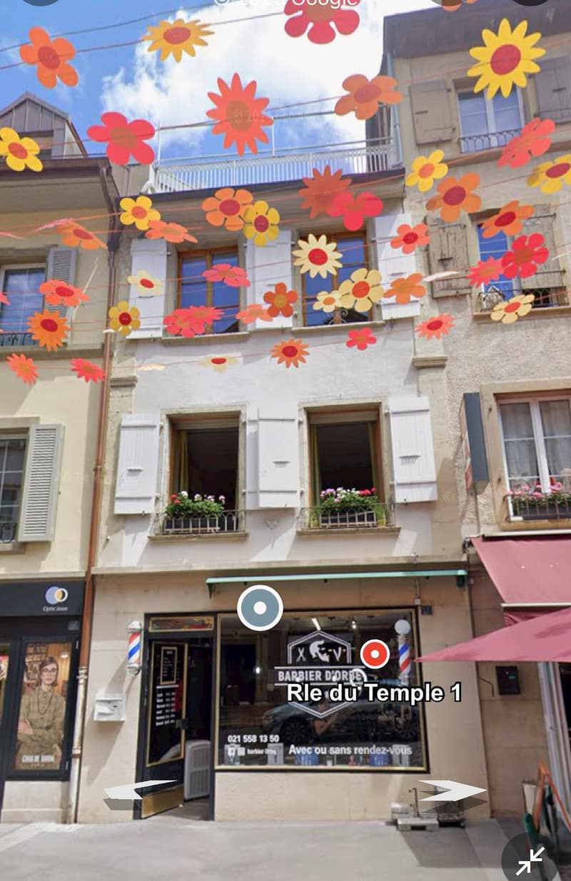 Front of a building with decorative flowers, barber shop, balcony with flower planters, street level with awnings, and a red awning on the right.
