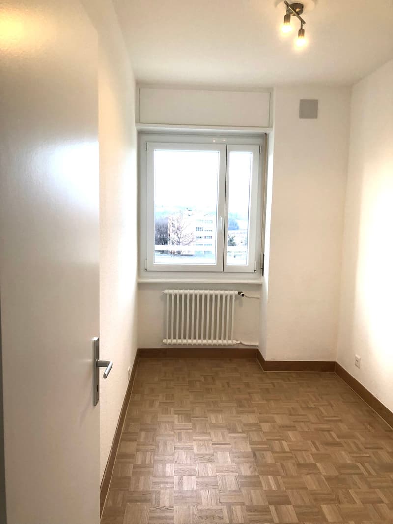 Empty room with white walls, wooden parquet floor, white radiator, large window, open door