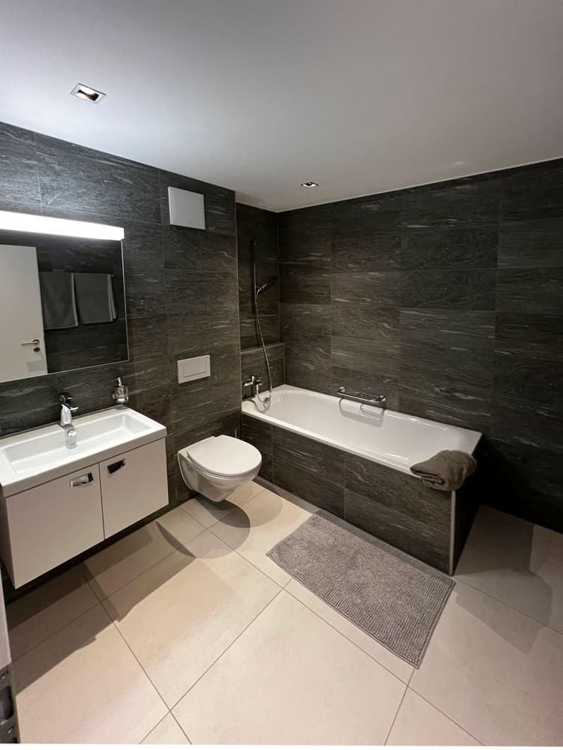 modern bathroom with white bathtub, grey tiles, white vanity with sink, toilet, wall-mounted mirror, under-cabinet lighting, towel rack, and towel.