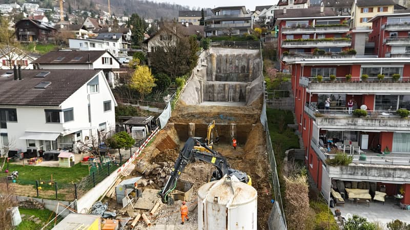 Houses, construction site, excavator, machinery vehicles, building in different stages of completion, green grass, people standing on balcony