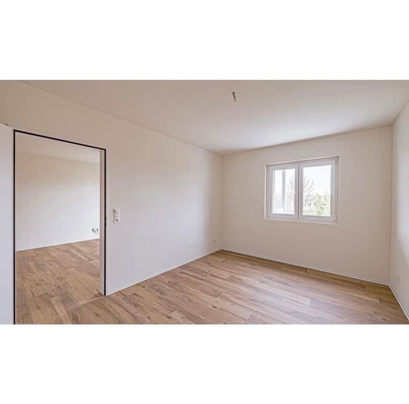Empty room with white walls, wooden floors, an open doorway leading to another room, and a window allowing natural light.