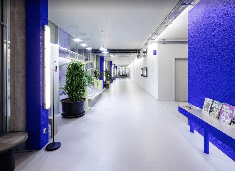 Long corridor with white tiles, blue painted walls, a bench, plants, and wall lamps.