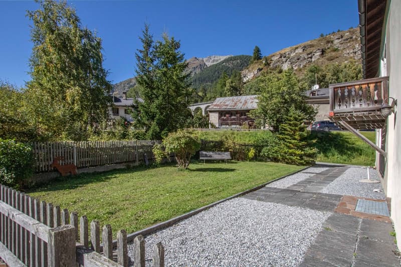 green grassy lawn, gravel path, bench, wooden fence, house in background, mountains
