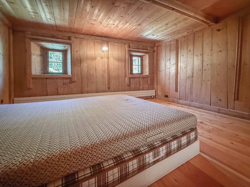 A bedroom with wooden paneled walls and floor, two small windows, a bed, and a wall-mounted lamp.