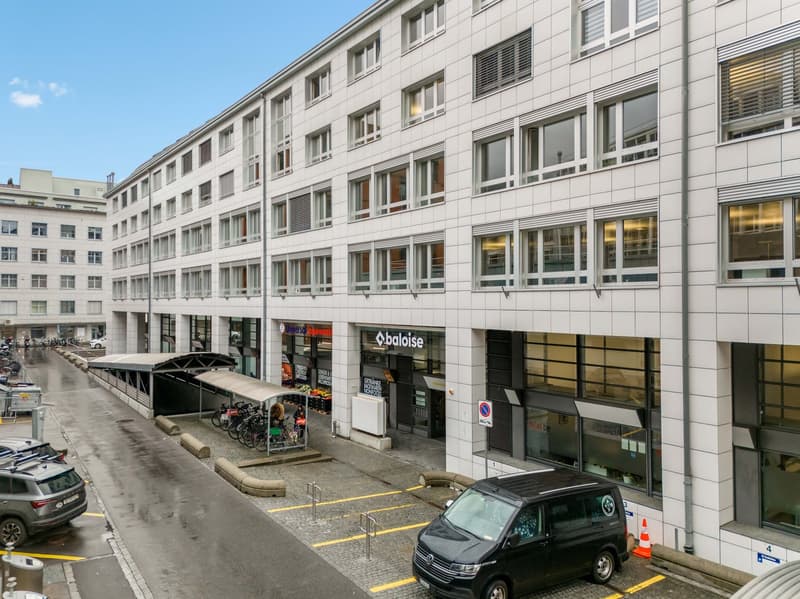 White building with multiple windows, ground floor with shops, parking area in front