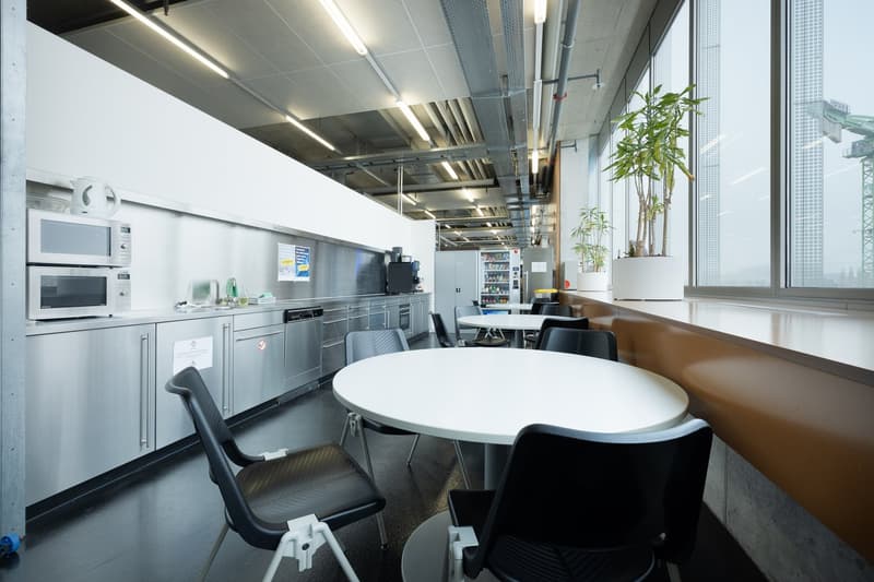 Contemporary open-plan workspace with round white tables, black chairs, steel counters, kitchen appliances, and large windows.