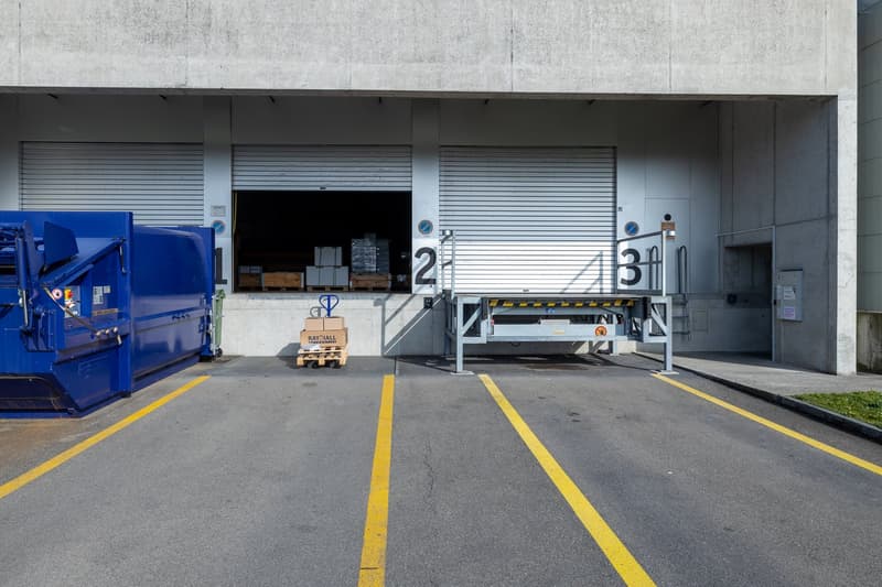 commercial building with large roll-off dumpster, large sliding door, metal lift bridge, yellow parking lines, two storage areas with pallets and boxes