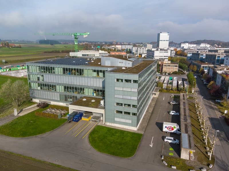 Large office building with glass walls, solar panels on the roof, spacious parking lot, multiple vehicles parked.