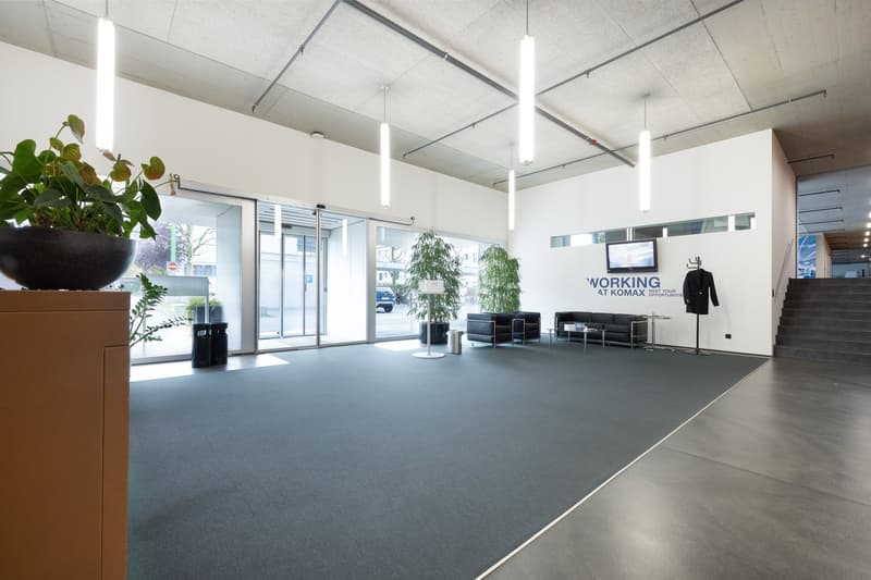 A modern, minimalist lobby with gray carpet, a wooden reception desk, large windows, and greenery. A wall features the text 'WORKING AT KOMAX'. Chairs, a couch, and a table are placed against the wall.