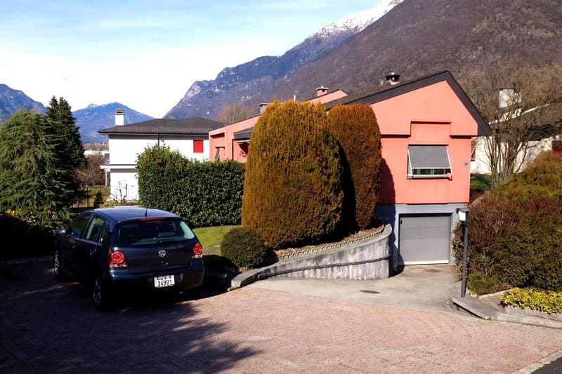 A two-story house painted in coral, located at the base of a mountain, features a gray garage, a small driveway, and a vehicle parked in front.