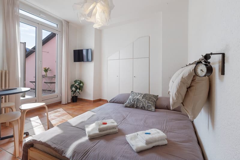 small bedroom with bed, bedside table, chairs, TV, sliding glass door, balcony, plant