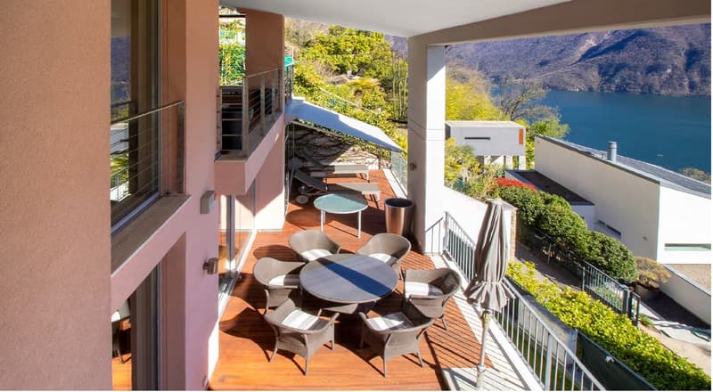 Outdoor terrace with wooden deck, round table, chairs, and a closed umbrella. View of a lake and mountains.
