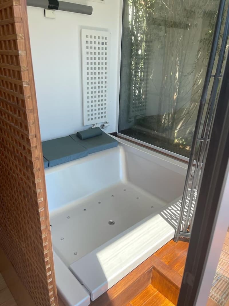 A modern bathroom with a large bathtub, a bench with cushions, a radiator, and a large window.