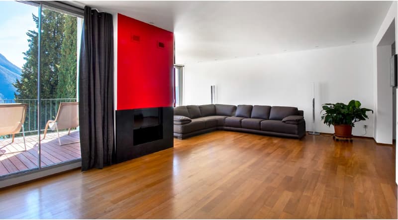 Spacious living room with hardwood floors, a red accent wall, a sectional sofa, and a fireplace. Large windows lead to a balcony.
