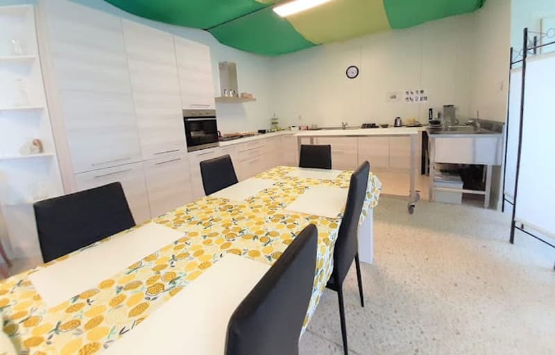Dining room: long table, chairs, white cabinets, white counter, kitchen with oven, stove, sink, refrigerator, coffee machine
