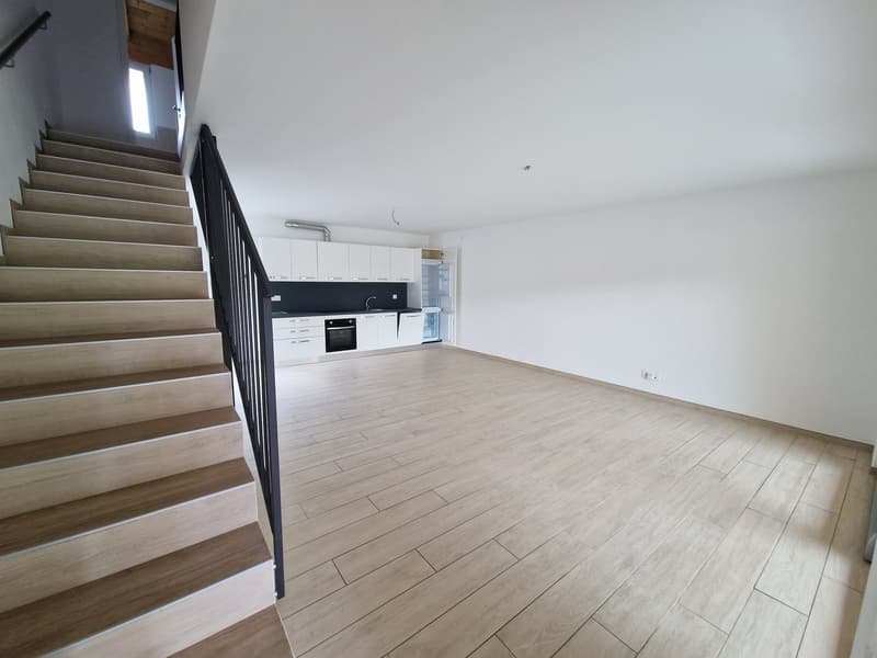 Empty room with wooden floor, white walls, wooden staircase, kitchen with white cabinets, black countertop, and appliances
