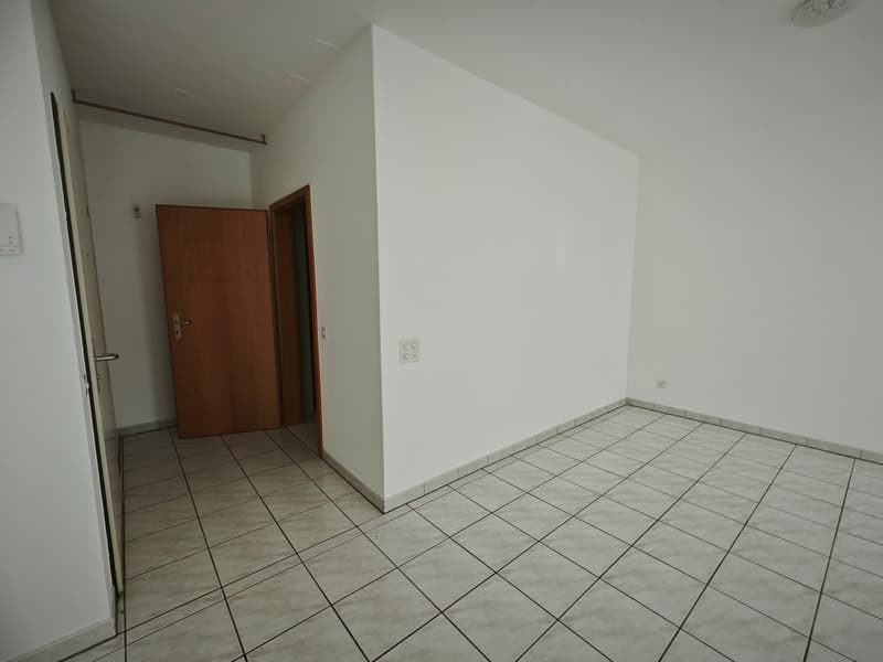 Empty room with white walls, tiled floor, wooden door, power outlets, and a light fixture on the ceiling.