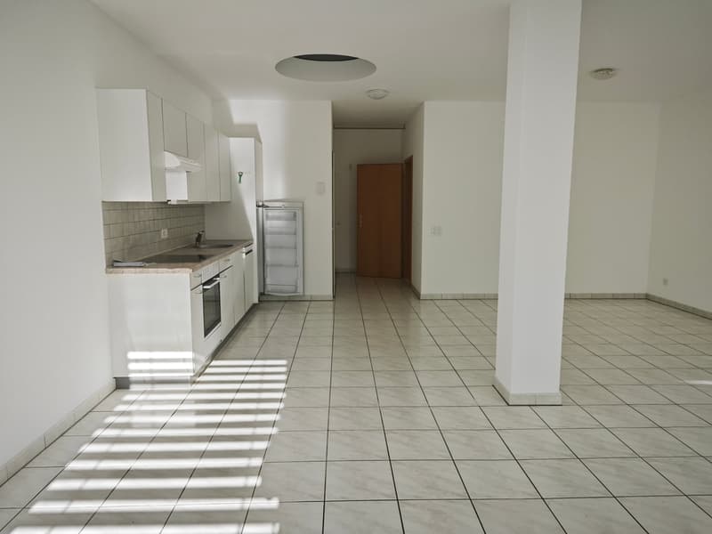 Empty kitchen with white tiles, white cabinets, sink, oven, refrigerator, and a door leading to another room.