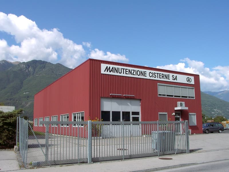 Red industrial building with large garage doors, multiple windows, and a sign reading 'MANUTENZIONE CISTERNE SA'.