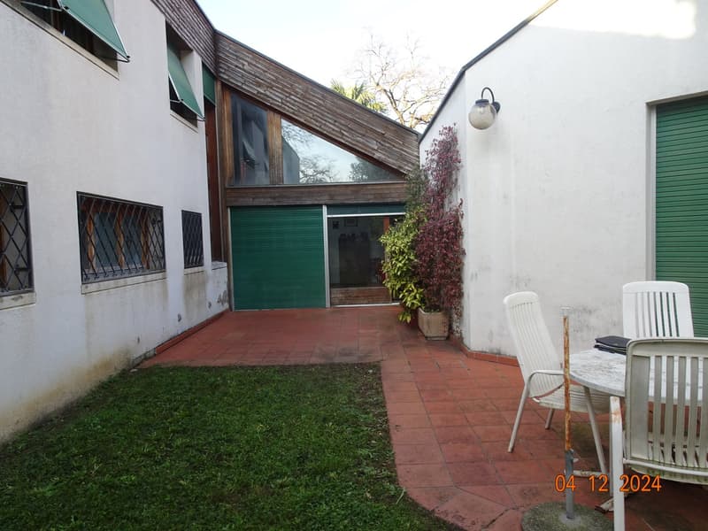 Red brick tiles garden with green plants, a green garage, white chairs, and a table