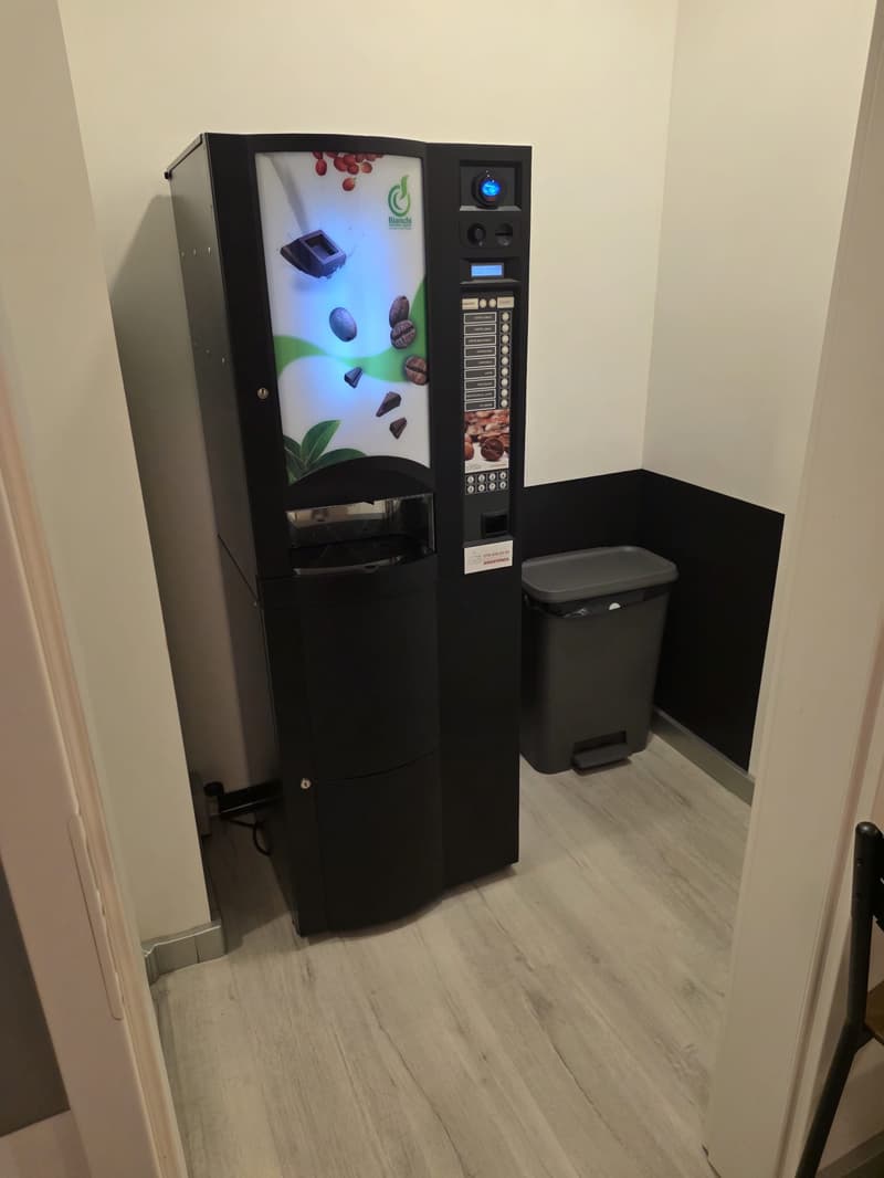 A black coffee vending machine in the corner with a trash can nearby. The wall is white, and there is a small table with a seat on the right side.