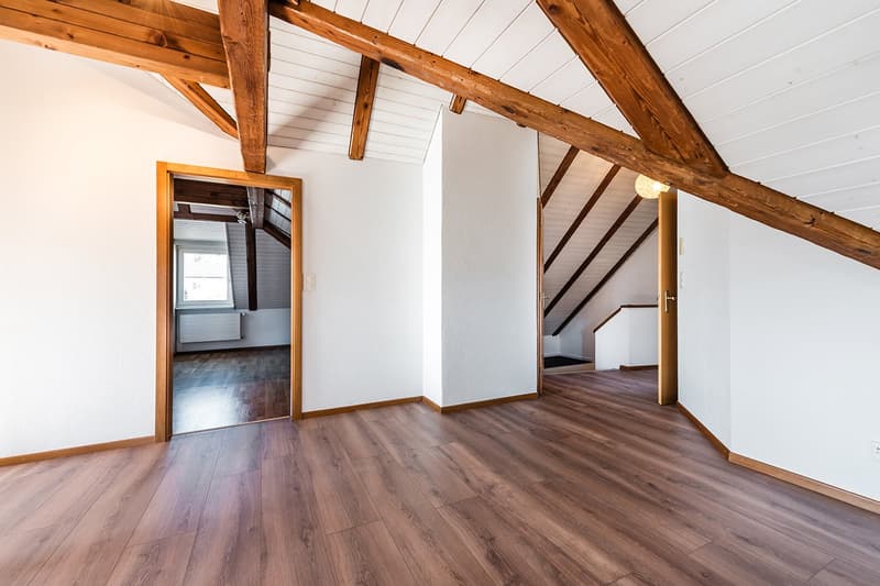 A wooden attic room with hardwood floors, wooden beams, white walls, an open door, and an empty room with a window.