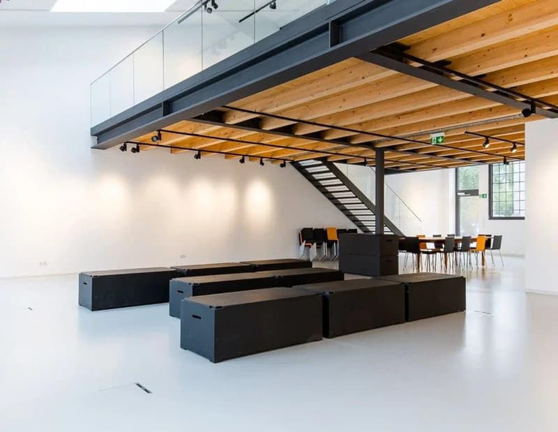 Open space with black storage boxes, metal ceiling beams, glass railing, white walls, wooden flooring on upper level, desk and chairs.
