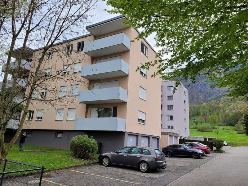3 story building, beige and gray, balconies, parking lot, cars parked, trees, mountains in the background