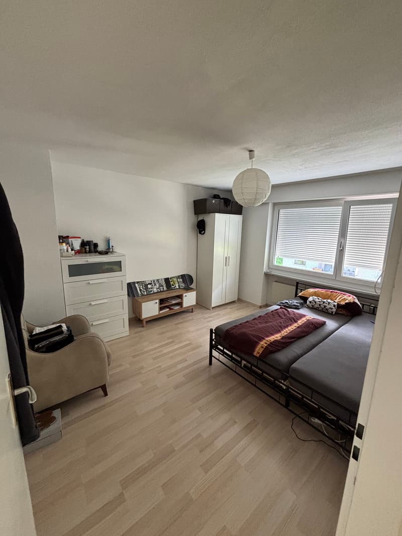 Bedroom with two beds, wooden floor, white walls, window with blinds, wardrobe, desk, chair, lamp, skateboard, and various items on the dresser.