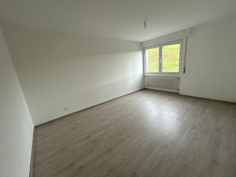 An empty room with a white wall and wooden floor. There is a window with a white radiator below. There are two white outlets on the wall.