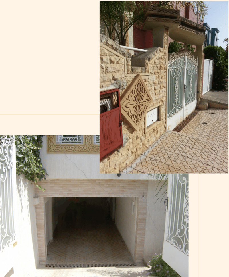 A stone building with a red gate, a patterned wall design, and a white brick wall. The entrance is paved with bricks, and there is a garage with a tiled floor.