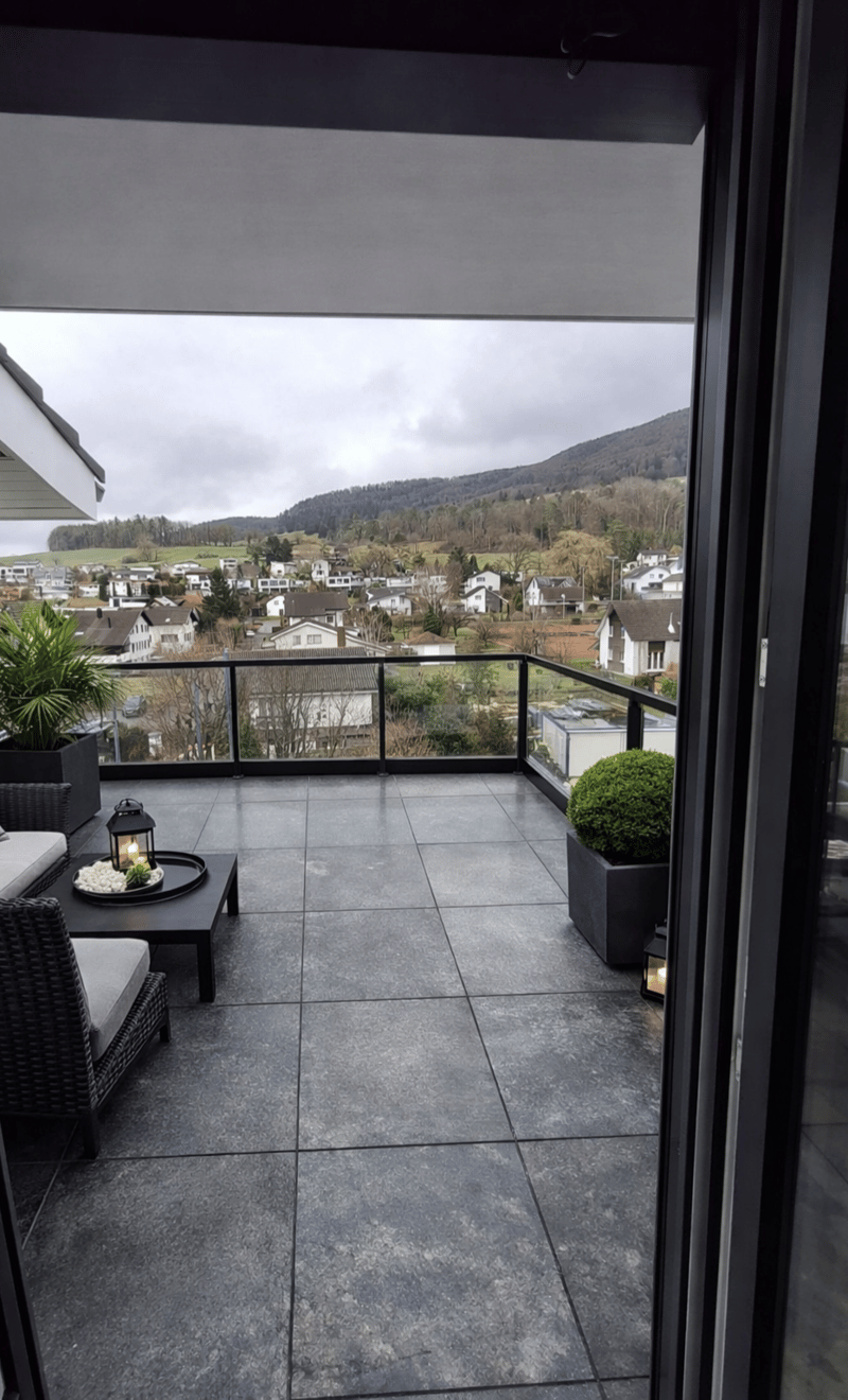 covered balcony with gray tiles, table with a lantern, wicker chairs, plant, metal railing, city view