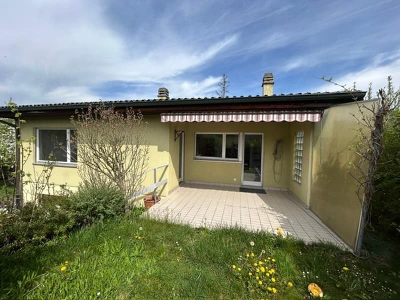 Single-story house, yellow exterior, tiled patio, red and white awning, glass windows, surrounded by grass and plants