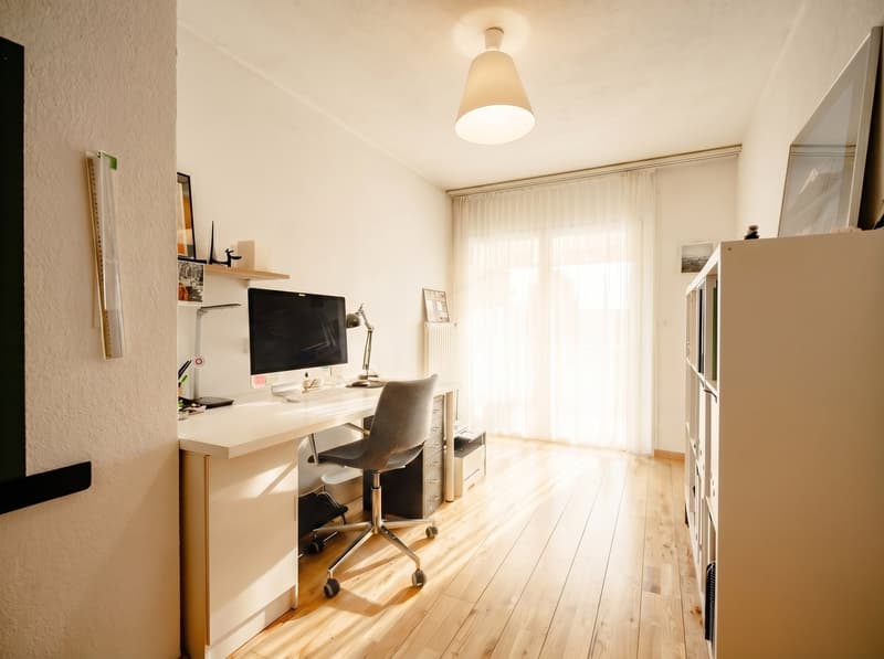 Desk with computer, chair, shelves, lamp, wooden floor, white walls, window with curtains