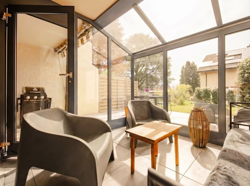 modern living room with glass walls, wooden table, gray chairs, bamboo vase, outdoor grill, garden view
