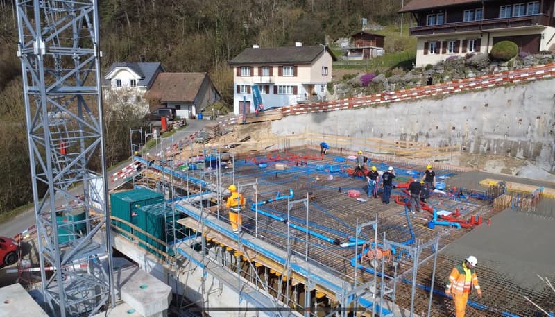 Under construction building, workers, scaffolding, pipes, concrete foundation, green containers, machinery, houses in the background