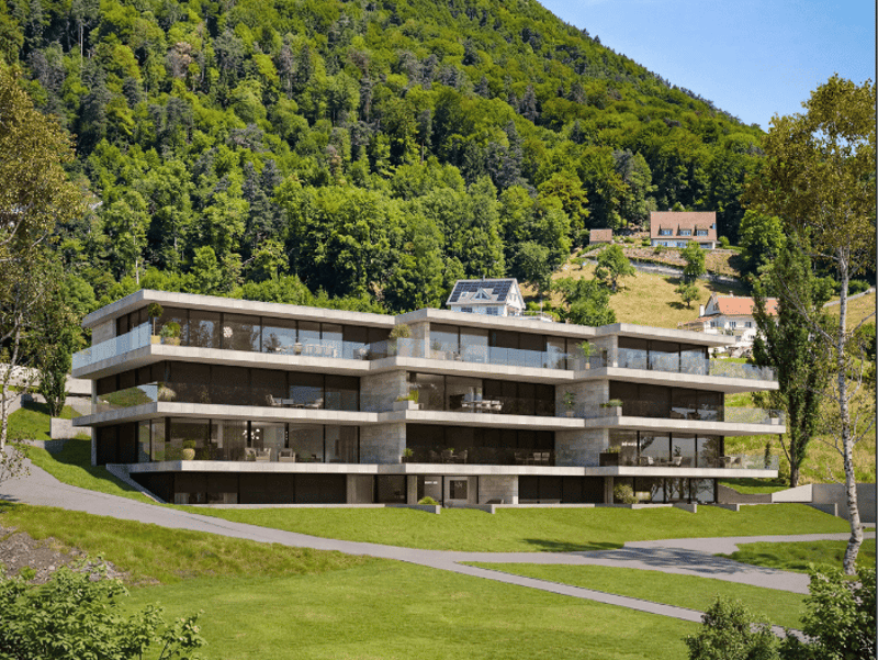 Modern multi-story building with glass walls, balconies, and solar panels on the roof, set in a lush green landscape with a mountain in the background.