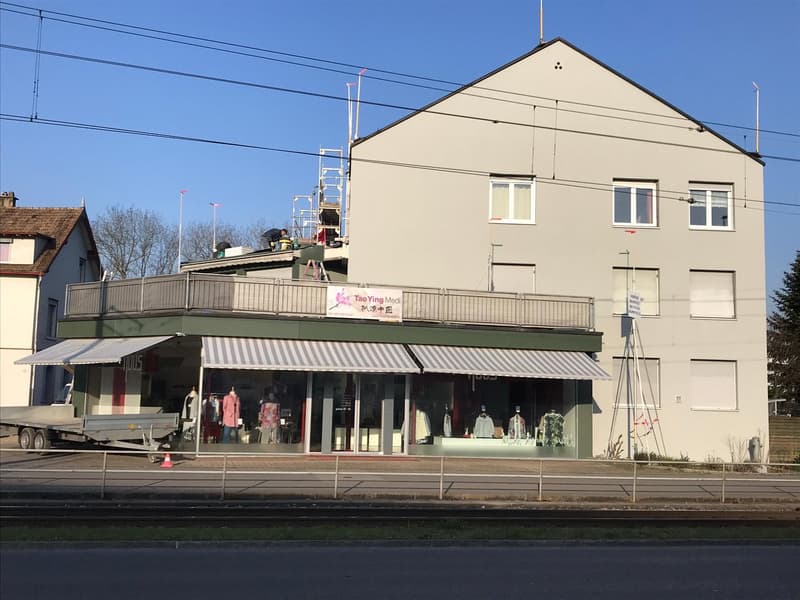 A 2-story building with gray walls, a tiled roof, and a metal awning, featuring multiple windows, an attached trailer, and workers performing repairs on the roof.
