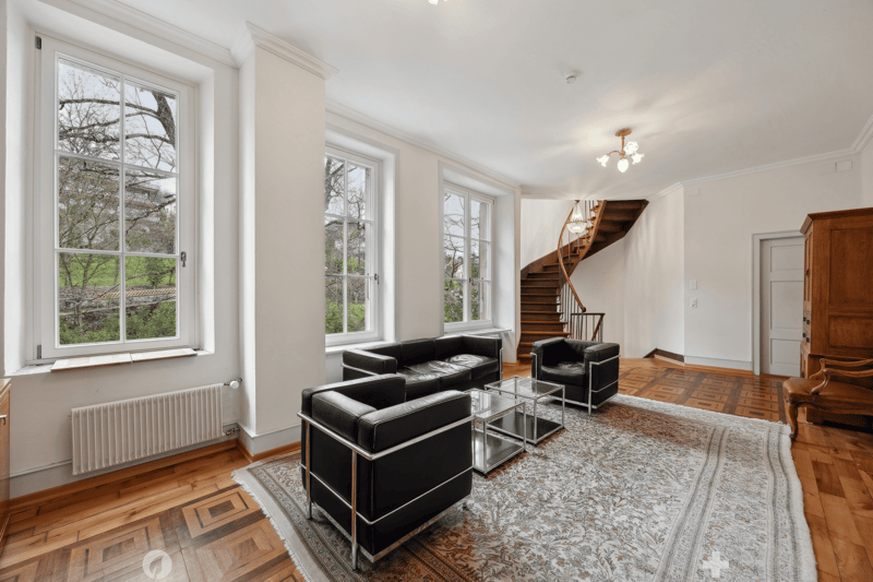 Open layout with two black leather sofas and glass tables, wooden floor, white walls, windows, staircase, chandeliers, radiator